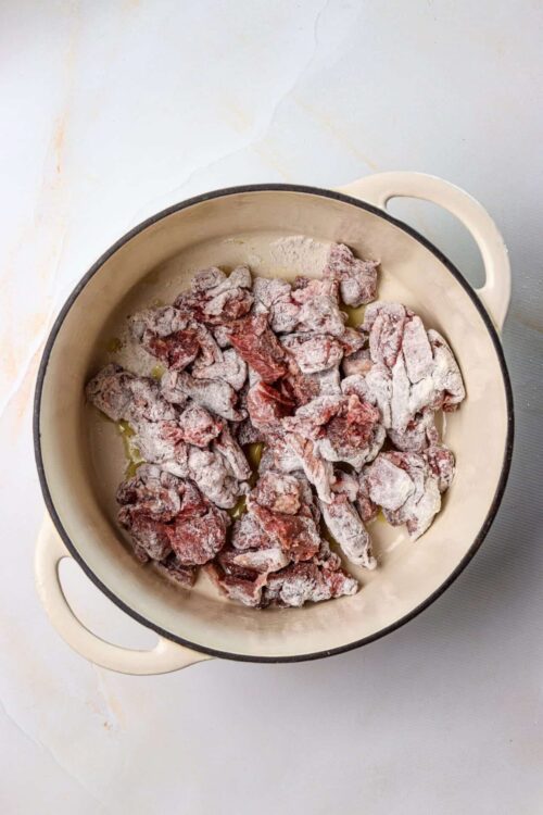 Cubed raw beef seasoned for stew in a white ceramic cooking pot.