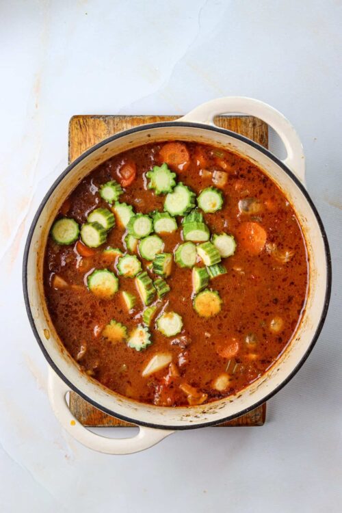Hearty vegetable soup with sliced cucumbers on top, in a white cast iron skillet, over a wooden cutting board.
