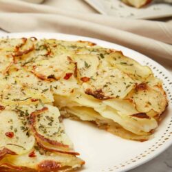 Buttered potato gratin with herbs on a white plate, baked potato slices topped with parsley.
