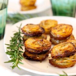 Baked eggplant stacks on a white plate, garnished with fresh rosemary and served with green glasses.