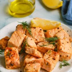 Juicy baked salmon cubes on a white plate with lemon wedges, dill garnish, and a glass of olive oil in the background.