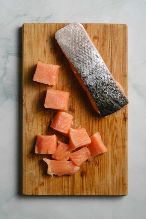 Fresh salmon fillet with skin on and cubed salmon pieces on a wooden cutting board for cooking or preparation.