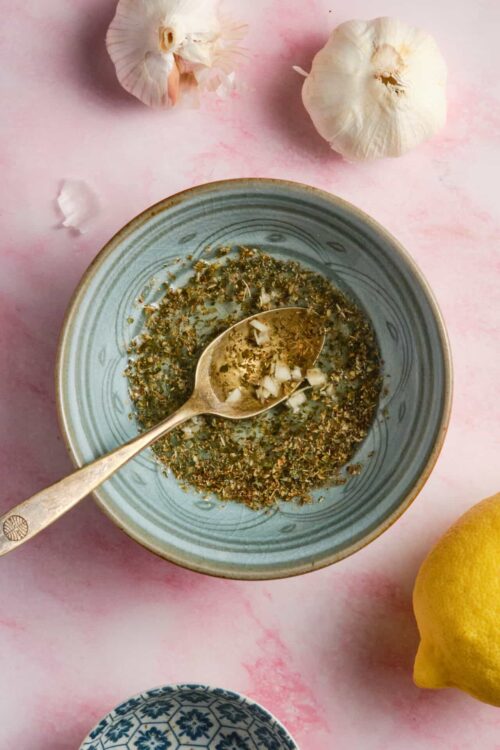 Dried garlic and herb seasoning mix in a blue bowl on pink background with garlic and lemon.
