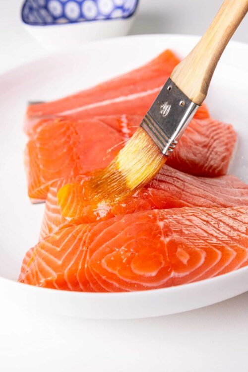 Fresh raw salmon being brushed with marinade on a white plate, ready for cooking or baking.