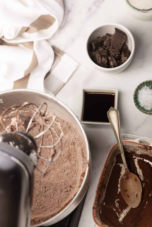 Rich chocolate cake batter in a stand mixer with chocolate chunks and baking ingredients in the background.