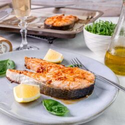 Pan-seared salmon with lemon wedges on a white plate, fresh basil and a fork, with a glass of white wine in the background.