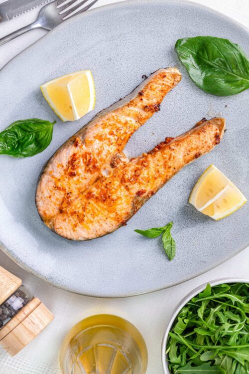 Salmon fillet with lemon wedges and fresh basil on a light gray plate.