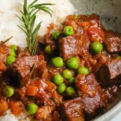Beef stew with green peas and fresh rosemary served over rice in a speckled bowl.