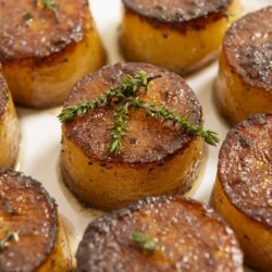 Golden caramelized scallops wrapped in bacon with fresh thyme garnish for savory seafood dish.