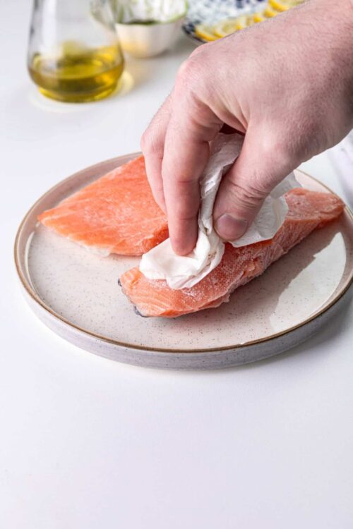 Fresh salmon fillet being seasoned with paper towel on a ceramic plate.