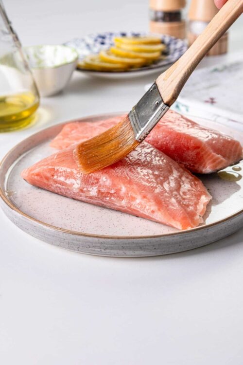 Raw salmon fillets being brushed with oil on a ceramic plate.