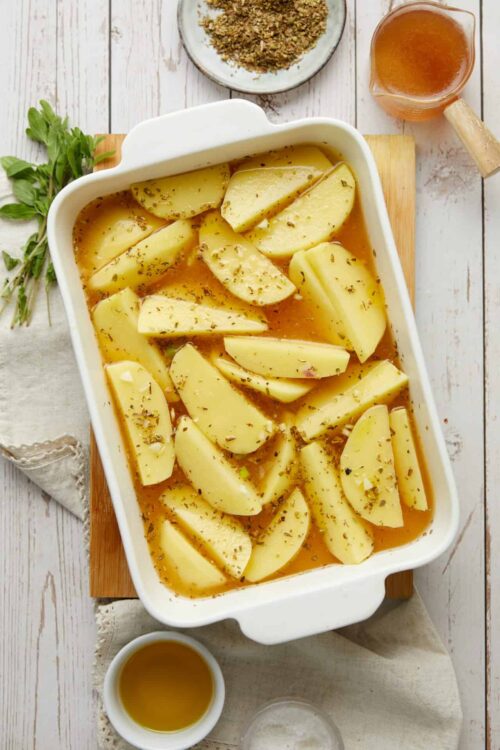 Baked potatoes with herbs and gravy in a baking dish on a white wooden surface.