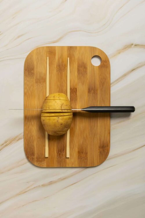 Sliced potato being prepared with a sharp knife on a wooden cutting board.