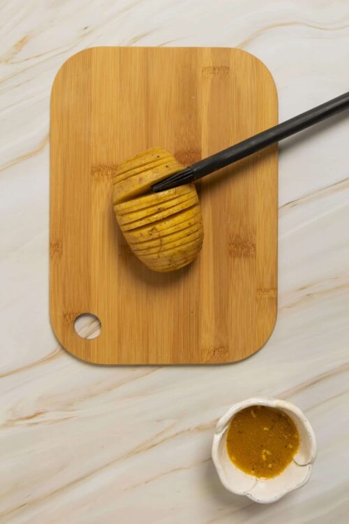 Sliced Hasselback potatoes on bamboo cutting board with basting brush and sauce bowl, perfect for baked potato recipes.