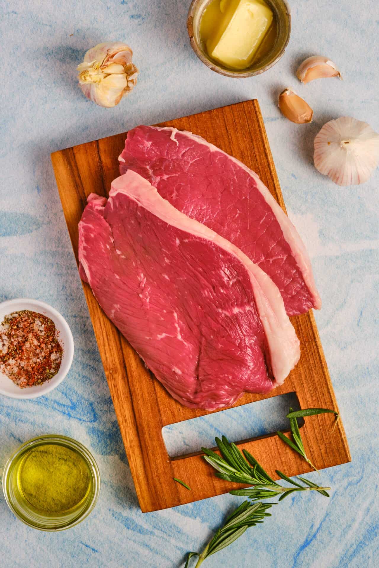 Raw beef steak on wooden cutting board with garlic, butter, herbs, and spices.