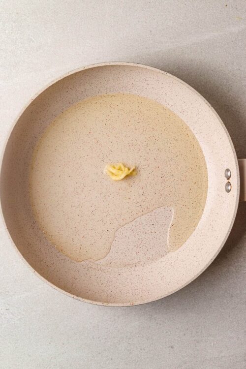 Butter melting in a beige non-stick frying pan with a speckled surface.