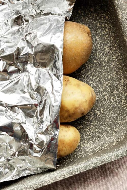 Baked potatoes partially wrapped in aluminum foil on a baking sheet with some salt scattered around.