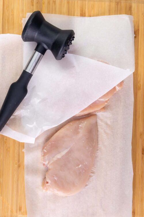 Hammer tenderizing raw chicken breast on parchment paper on wooden surface.