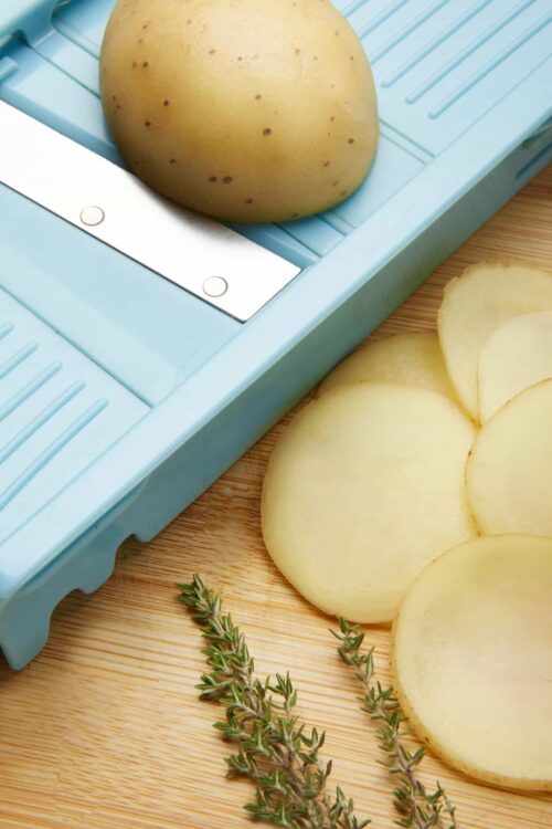 Fresh whole potatoes on a blue food dicer, sliced potatoes, and thyme on a wooden surface.