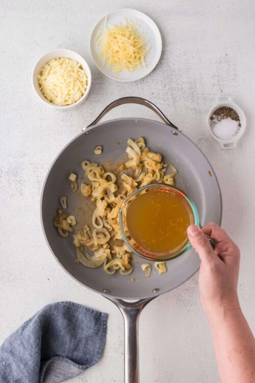 Creamy onion and cheese filling being prepared in a skillet with added chicken broth, surrounded by shredded cheese, salt, and pepper on a white countertop.