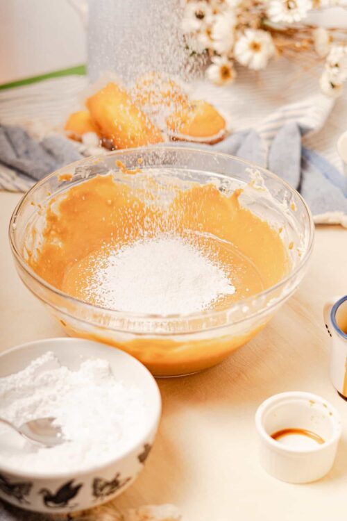 Flour being sprinkled into a mixing bowl with cake batter on a baking station.