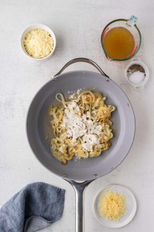 Creamy sautéed onions in a gray skillet with grated cheese and seasonings, ingredients ready for cooking.