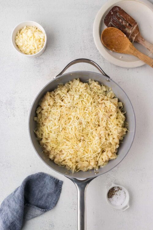 Creamy macaroni and cheese dish with shredded cheese in a stainless steel skillet, topped with cheese and ready for baking.