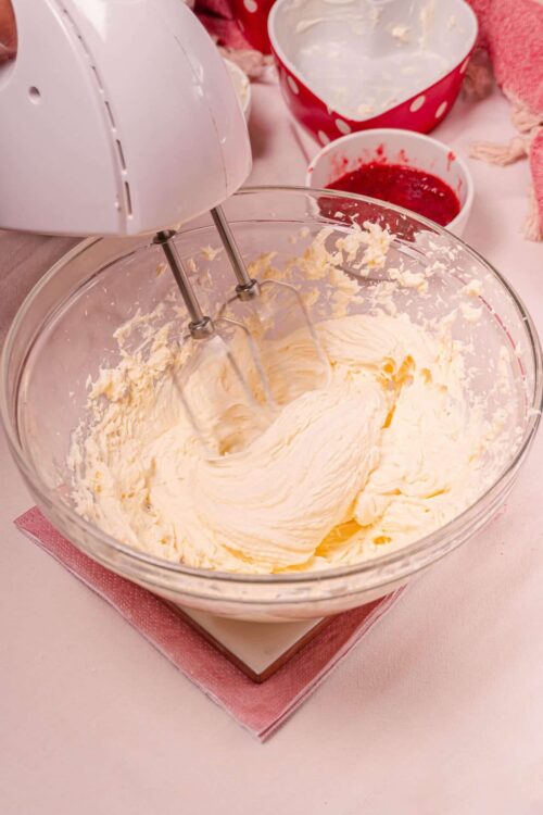 Fluffy buttercream frosting being whipped in a glass mixing bowl using an electric hand mixer.