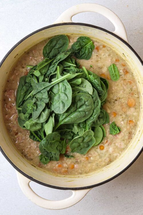Fresh spinach added to creamy vegetable soup in a white enamel pot.