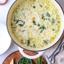 Savory vegetable chowder in a white pot with fresh herbs and cheese on a wooden cutting board.
