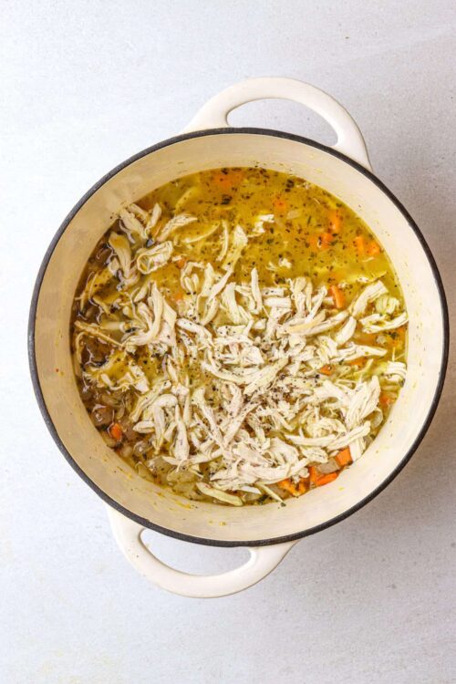 Homemade chicken soup with shredded chicken, vegetables, and herbs in a white enamel pot.