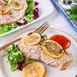 Lemon herb baked salmon fillet with fresh herbs and citrus slices on a white plate.