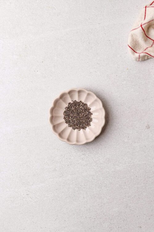 Raw chia seeds in a scalloped ceramic dish on a light surface, minimal food photography for healthy eating or baking.