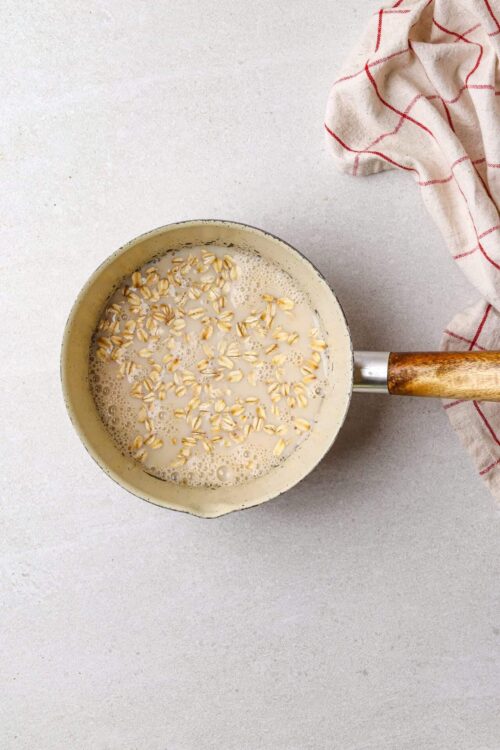 Creamy oat milk in a small saucepan with oats, on a light countertop with a red-checkered cloth.