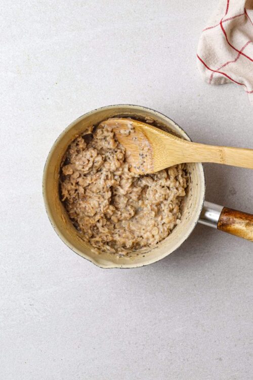 Creamy oatmeal with black pepper in a beige saucepan with wooden spoon.