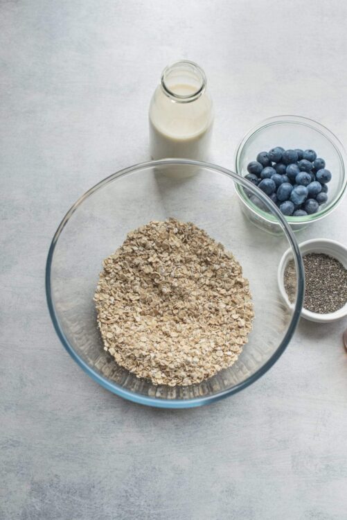 Oatmeal in a clear glass bowl on a white surface, with a bottle of milk, blueberries, chia seeds, and more oats around it.