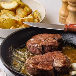 Crispy roasted potatoes with herbs in a white bowl, served near cast iron skillet with cooked steaks on a white surface.