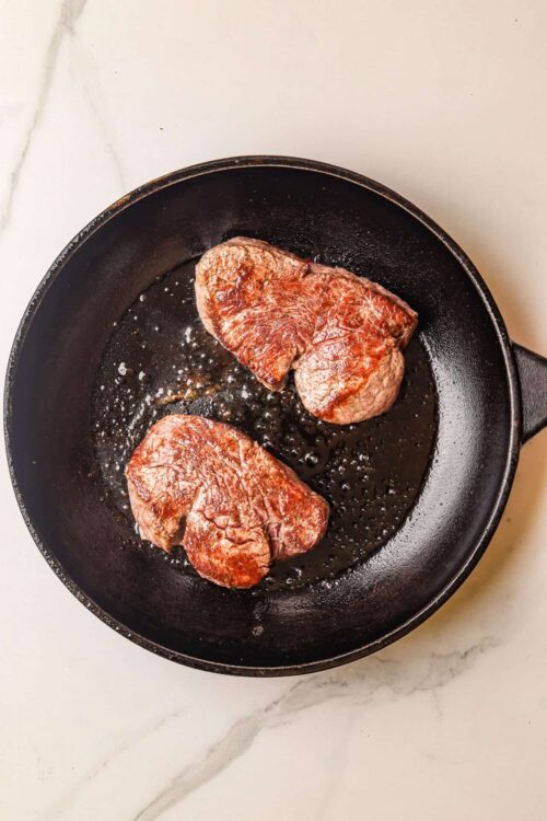 Sirloin steaks cooking in a black cast iron skillet on white marble countertop.