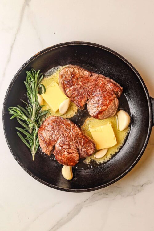 Juicy seared steaks with garlic, butter, and fresh rosemary in a black skillet.