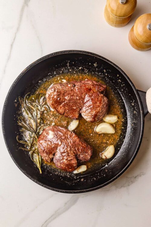 Juicy seared steaks with garlic, rosemary, and herbs in a cast iron skillet for flavorful cooking.