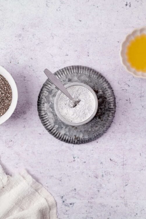 Creamy chia seed pudding in a glass jar with a silver spoon on a textured surface.