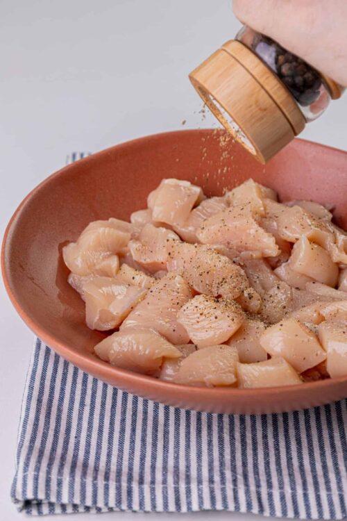 Raw chicken breast pieces seasoned with black pepper in a pink bowl, with a hand sprinkles spice over the chicken.