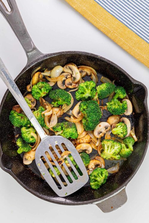 Steamed broccoli, mushrooms, and onions cooking in a black skillet with a metal spatula.