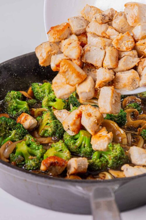Cooked chicken being added to a skillet with broccoli and mushrooms for a healthy stir-fry recipe.