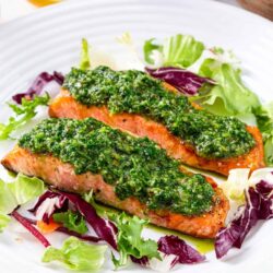 Salmon with green herb sauce served over mixed greens salad on white plate.