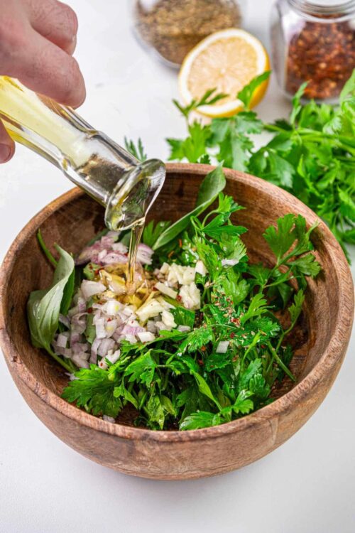 Fresh herb salad with olive oil being poured, ingredients include parsley, basil, onion, and garlic.