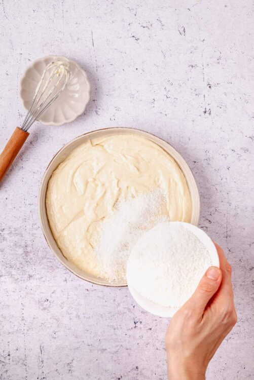 Creamy homemade cake batter with flour being added, on textured white surface, ideal for baking recipes.