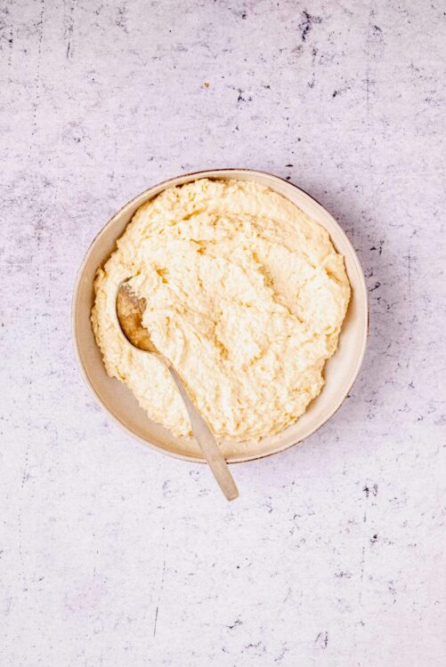 Creamy mashed potatoes in a beige bowl with a spoon, on a textured light gray surface.