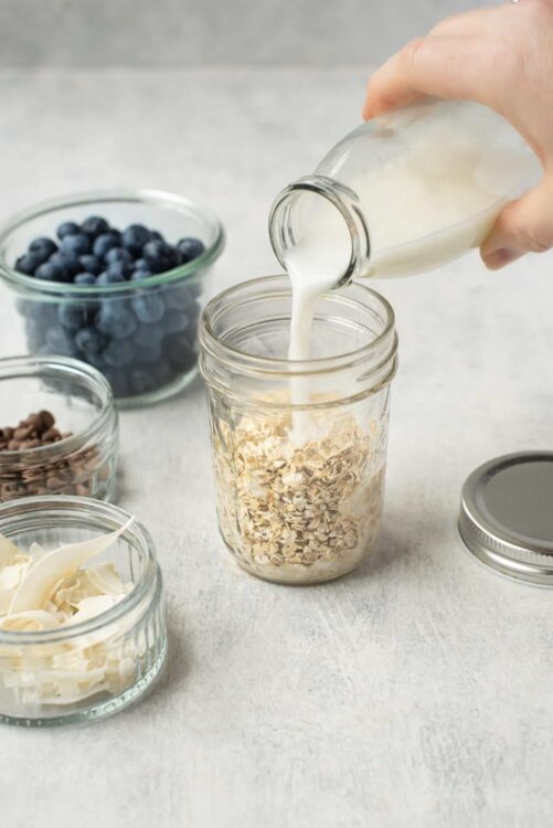 Cream being poured into a jar of oats with blueberries, chocolate chips, and white chocolate shavings on a light background.