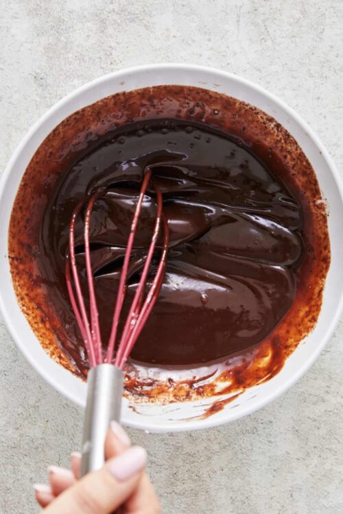 Rich melted chocolate being mixed with a hand whisk in a white bowl.